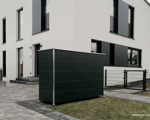 . The image shows a modern residential building with a minimalist design. There is a dark-colored storage unit in the foreground, placed on a paved area next to the house. The building has large windows and a flat exterior, with a small fence and gate visible near the entrance. The landscape includes a grass lawn and some paving stones.