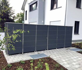 . The image shows a modern, outdoor storage unit or bin designed for use in a garden or yard. It has a sleek, structured look with a dark finish and is situated against a paved path and landscaped area with mulch and small plants. The unit is located beside a contemporary house with white and gray walls.