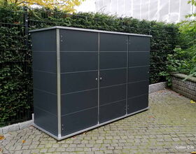 . Ein moderner, dunkler Geräteschrank steht auf einem gepflasterten Bereich im Garten, umgeben von grüner Hecke.