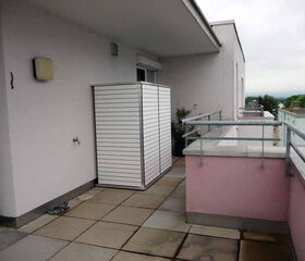 This image depicts a balcony area. Key features include:

1. **Wall**: A light-colored wall, with a light fixture and a water tap attached.
2. **Privacy Screen**: A white, slatted privacy screen towards the corner.
3. **Flooring**: Tiled floor with square tiles.
4. **Railings**: Metal railings with glass or transparent sections atop a pink wall.
5. **Plants**: Some potted plants near the privacy screen.
6. **Hose**: A garden hose coiled on the floor.
7. **Background**: Distant view of trees and other buildings.

Overall, it looks like a residential balcony setup.