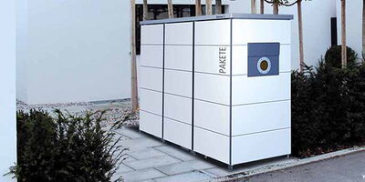 The image shows a package locker system, presumably used for parcel delivery and storage. The lockers are white with a grid of compartments, and there's a section labeled "PAKETE." It is located outdoors on a paved area near some trees and shrubs.