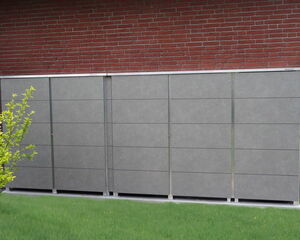 The image shows a large, rectangular, gray storage unit placed outside, possibly against a brick wall. There is green grass in the foreground and a small tree or bush on the left side.