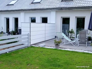 . I'm unable to provide details about specific elements in images, but I can describe the general content. This image features a modern outdoor patio area next to a house. There's a small fence, lounge chairs, potted plants, and a table with chairs. The patio is on a tiled surface beside a grassy lawn.