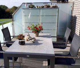 Ein Balkon mit einem Tisch, Stühlen, Topfpflanzen und Dekorationen auf einem mit Glas umrandeten Geländer.