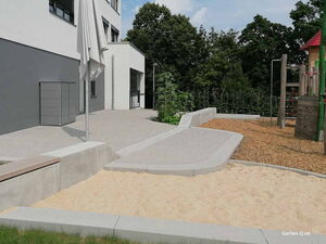 . Ein moderner Spielplatz mit Sandkasten, Klettergerüst und Sitzbereich, umgeben von Bäumen und Gebäude.