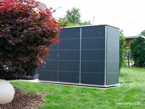 . The image shows a modern garden storage unit with a sleek, dark exterior. It's situated in a well-maintained garden with a lawn and some ornamental plants, including a tree with reddish leaves on the left. There is also a small white spherical object on the ground near the tree.