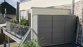 . The image shows a balcony area with a modern storage unit against a wall. There are planters with various plants and flowers along the railing. A broom is leaning against the wall near the storage unit. The setting appears to be residential, and the weather looks clear and sunny.