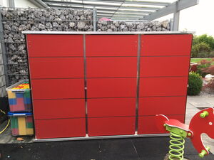 . The image depicts a backyard or garden area with several objects:

1. A large red cabinet or storage unit with metal trim and horizontal lines, likely for outdoor storage.
2. A transparent plastic box containing colorful blocks or toys, possibly for children.
3. A metal wire mesh filled with rocks, functioning as a gabion wall or fence.
4. A child’s spring rider playground toy with a seat shaped like a red animal, on a green spring.
5. Part of a garden in the background with plants and a pathway.

The setting appears to be a modern and organized outdoor area, possibly for a residential space.