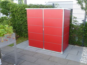 . The image features a red outdoor storage shed placed on a patio. The shed is surrounded by a neatly landscaped area with green shrubs in the background. To the left, there's a raised metal planter with strawberries growing in it. The area is paved with grey tiles.