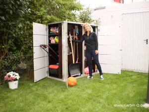 . The image shows a person standing next to an open garden storage unit. Inside the storage, there are various items such as gardening tools, a snow shovel, a rake, some sports equipment like balls, and possibly other outdoor items. There are flowers in pots nearby, and the setting is an outdoor garden or backyard area.