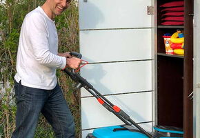 . The image shows a person pushing a blue lawnmower toward a storage cabinet. The cabinet has shelves with neatly stacked red cushions and some colorful items. It's set in an outdoor area with greenery around.