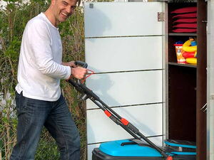 . The image shows a man smiling and pushing a lawn mower. He is standing on grass next to a storage shed. The shed has shelves with some items, including what appear to be red cushions and colorful toys. The setting is outdoors, adjacent to a building.