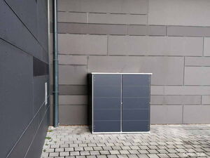 . Ein grauer Schrank steht auf einem gepflasterten Boden an einer modern gestalteten, grauen Wand.