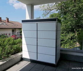 . The image shows a modern outdoor storage cabinet or shed situated on a patio or balcony. It's sleek and modular in design with a white finish and dark trim. There is greenery and a residential building in the background.