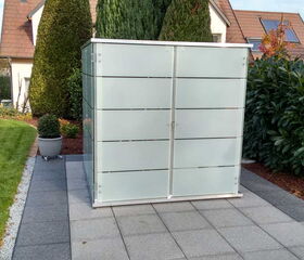 . The image depicts an outdoor scene with a modern, frosted glass enclosure on a patio area. The enclosure likely serves as a storage unit or utility cabinet. It is situated on a tiled surface with neatly manicured grass and shrubs surrounding it, and a house is visible in the background.