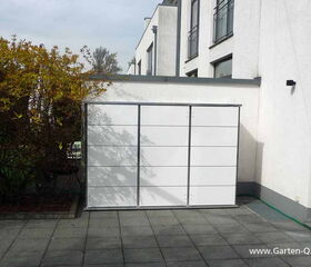. The image shows a white storage unit or cabinet placed outdoors on a patio, next to a building with multiple windows. There are trees or shrubs on the left side, and the ground is tiled with large square tiles. The website "www.Garten-Q.de" is visible in the image.