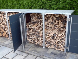 . The image shows a modern storage unit filled with stacked firewood. The unit has multiple compartments with open doors, allowing access to the wood. It's positioned on a paved surface, and there's greenery in the background.