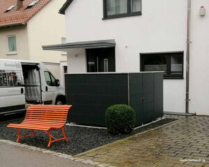 The image shows a modern residential setting. Key elements include:

1. A white house with black-trimmed windows.
2. A black structure or enclosure next to the house.
3. An orange bench made of metal slats.
4. A round green shrub in front of the black enclosure.
5. A paved pathway leading to the house.
6. A vehicle parked on the left side with "Garten Q" branding and images related to gardening or storage solutions.

There are also neighboring houses in the background.
