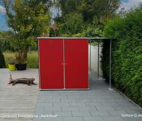 Ein rotes Gartenhaus mit einer Eidechse davor auf einer Terrasse, umgeben von Pflanzen.