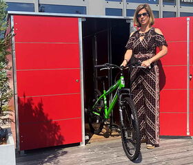 . Frau mit Sonnenbrille holt ein grünes Fahrrad aus einem roten Schuppen.