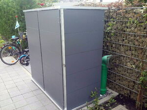 . The image shows a sleek, modern metal or plastic enclosure, possibly for a utility like a heat pump or air conditioning unit, situated in a garden area. There's a bicycle with a child seat nearby, a stone-filled wire fence providing privacy, and some greenery around the structure. The enclosure has a green ventilation pipe with a protective grill.