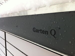 . The image shows a close-up of a structure with water droplets on its surface. The text "Garten[Q]" is visible, and there is snow on top of the structure. It appears to be part of a modern outdoor or garden storage unit.