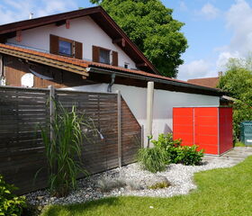 Ein Haus mit Holzzaun, Garten mit Kies und Pflanzen, rotes Schuppen, großer Baum im Hintergrund, blauer Himmel.