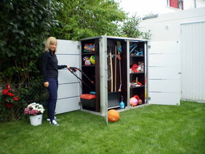 . The image shows a woman standing next to an open outdoor storage cabinet in a garden setting. The cabinet contains various items, including:

1. Gardening tools (rake, shovel, etc.)
2. A variety of balls (basketball, soccer ball)
3. A lawn mower
4. Cleaning supplies
5. Buckets of flowers
6. Miscellaneous small items (spray bottle, containers)

The scene is in a neatly kept garden area with green grass and some foliage and flowers.