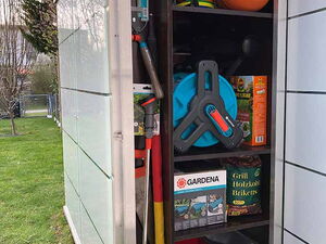. This image shows the contents of an outdoor storage cabinet. Inside, you can see:

1. Various gardening tools and equipment.
2. A basketball and some brightly colored juggling balls.
3. A hose reel.
4. Boxes labeled "Gardena" with gardening supplies.
5. A bag of grill charcoal briquettes.
6. Other items like a saw and small boxes, possibly for storage or organization.