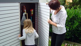 . The image shows two people, an adult and a child, opening a garden storage shed. The shed contains tools like a rake and a saw. It's set in a yard with plants and a lawn visible around it.