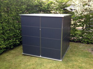 . The image shows a modern, dark-colored garden shed placed on a grassy lawn. It has a sleek design with clean lines, and is set against a backdrop of green foliage. This type of shed is often used for storing garden tools, equipment, or other outdoor items.