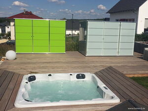 . The image shows a modern outdoor setup including a wooden deck with a built-in hot tub or jacuzzi. In the background, there are two storage units or cabinets, one bright green and the other in a pale color. The setting appears to be a backyard or garden area of a residential property, with houses visible in the background under a clear sky.