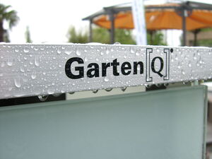 . The image shows a close-up of a metallic surface with water droplets on it. There is text on the surface that reads "Garten[Q]". In the background, there appears to be an outdoor structure with an orange canopy. The focus is mainly on the text and the water droplets.