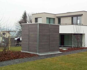 . The image shows a modern house with a cube-shaped structure in front of it. The cube appears to be covered with a wood-like material and has a metallic frame. The house has a flat roof, large windows, and is surrounded by a garden with sparse trees and grass. The setting appears to be residential, with overcast weather conditions.