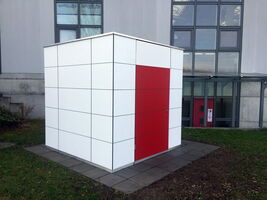 The image shows a cube-like structure with a red door, positioned on a tiled area next to a building. The structure is composed of white panels with a grid pattern, and it's located on grass near a building with large windows and a concrete wall.