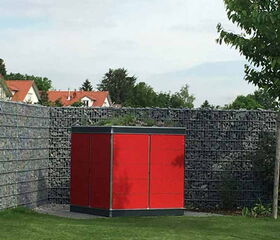 . Ein roter Gartencontainer steht auf einer grünen Rasenfläche, umgeben von einem Steingabionen-Zaun.
