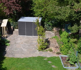 . The image shows a garden scene with a paved patio area and various plants and trees. There is a small structure that appears to be a shed or storage unit. Surrounding the patio are lush green plants, a vegetable garden, and a tree providing some shade. The area is well-maintained, with grass and decorative landscaping elements.