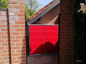 . Ein rotes Gartentor zwischen zwei Ziegelwänden, teilweise im Schatten.