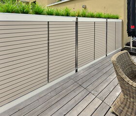 . The image shows a modern outdoor space with a focus on a privacy fence made of horizontal slats. Above the fence are planters with green plants. The area has a wooden deck with outdoor furniture, including a woven chair and a table with a closed umbrella. The setting suggests a patio or terrace.