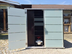 . The image shows a storage unit or cabinet with its doors open. Inside the cabinet, there are shelves and a small toy car visible at the bottom. The setting appears to be outdoors, possibly in a garden or patio area.