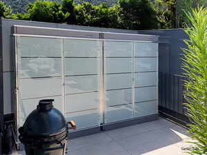 . Ein Grill und ein moderner Gartenzaun mit Blick auf Terrasse und grüne Bäume im Hintergrund.