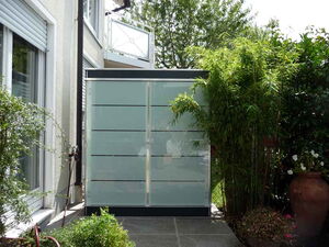 . This image shows a modern outdoor shower area with frosted glass panels for privacy. It is situated next to a house, surrounded by plants including bamboo, and has a tiled floor. There are pots and greenery around the shower, creating a lush and private space.
