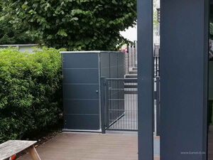 . The image shows an outdoor area with a modern garden storage unit or cabinet. It is situated next to a wooden deck and is surrounded by greenery, including a hedge and trees. There is a gate in front of the storage unit, and the materials used appear to be sleek and modern.