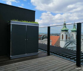 . Dachterrasse mit schwarzem Schrank, Glasgeländer, Blick auf historische Gebäude und bewölkten Himmel.