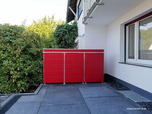 . The image shows a modern red storage unit placed outdoors on a patio or driveway. It has a sleek, minimalist design with multiple compartments and metallic accents. The unit is situated next to a house, complemented by a wall and window. Surrounding greenery, like bushes and trees, is visible in the background.