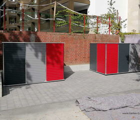 . The image shows two storage units with a modern design positioned on a paved area. Both units have a combination of dark gray, light gray, and red panels. They are placed in front of a brick wall, and there's a large structure with netting and plants behind the wall. A piece of fabric or covering material is laid out on the ground in front of the units.