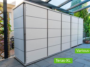 . Ein moderner, weißer Geräteschrank auf einer Terrasse mit grünem Pflanzenhintergrund.