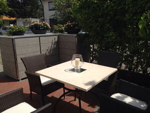 . The image shows an outdoor patio setup with a square table and four wicker chairs. The table has a light-colored surface with a glass candle holder that contains a candle. In the background, there are lush green bushes and planters with flowers on a wooden or composite structure. The setting seems to be a cozy outdoor seating area, possibly in a garden or a backyard.