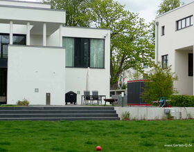 The image depicts a modern residential outdoor area with the following elements:

1. **Building**: Two modern white buildings with large windows.
2. **Grass Lawn**: A well-maintained grassy area.
3. **Outdoor Furniture**: A table with chairs and an umbrella.
4. **Barbecue Grill**: Located on the patio.
5. **Children’s Play Equipment**: Some play items are visible.
6. **Potted Plants and Bushes**: Various green plants around the area.
7. **Steps**: Leading from the patio to the lawn.

The setting appears to be a residential backyard or garden space.