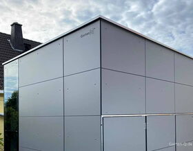 Modernes Gartenhäuschen mit Metallverkleidung neben einem Haus mit Schornstein.